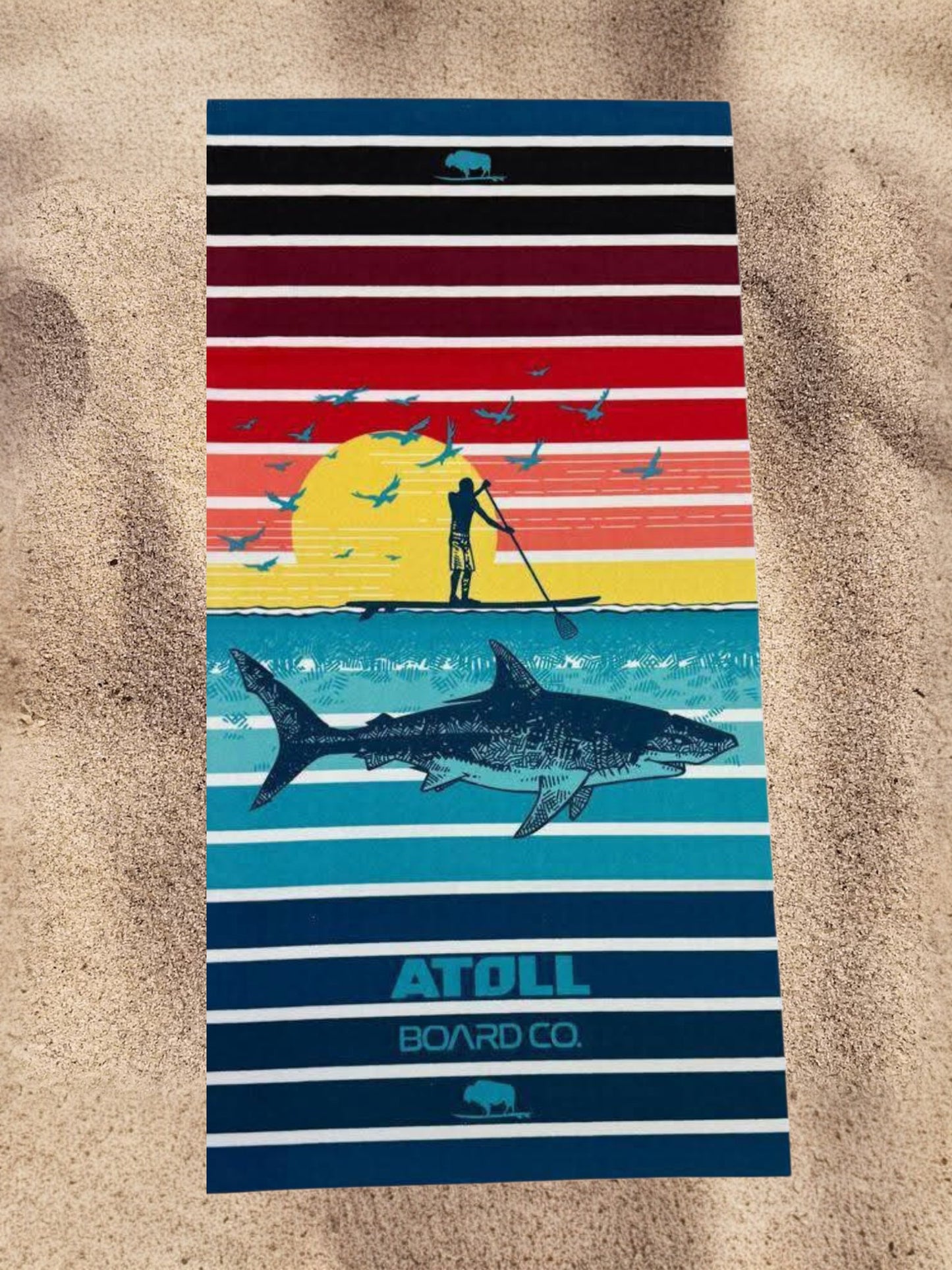 Striped towel with beach scene design and 'Atoll Board Co' branding on sand background