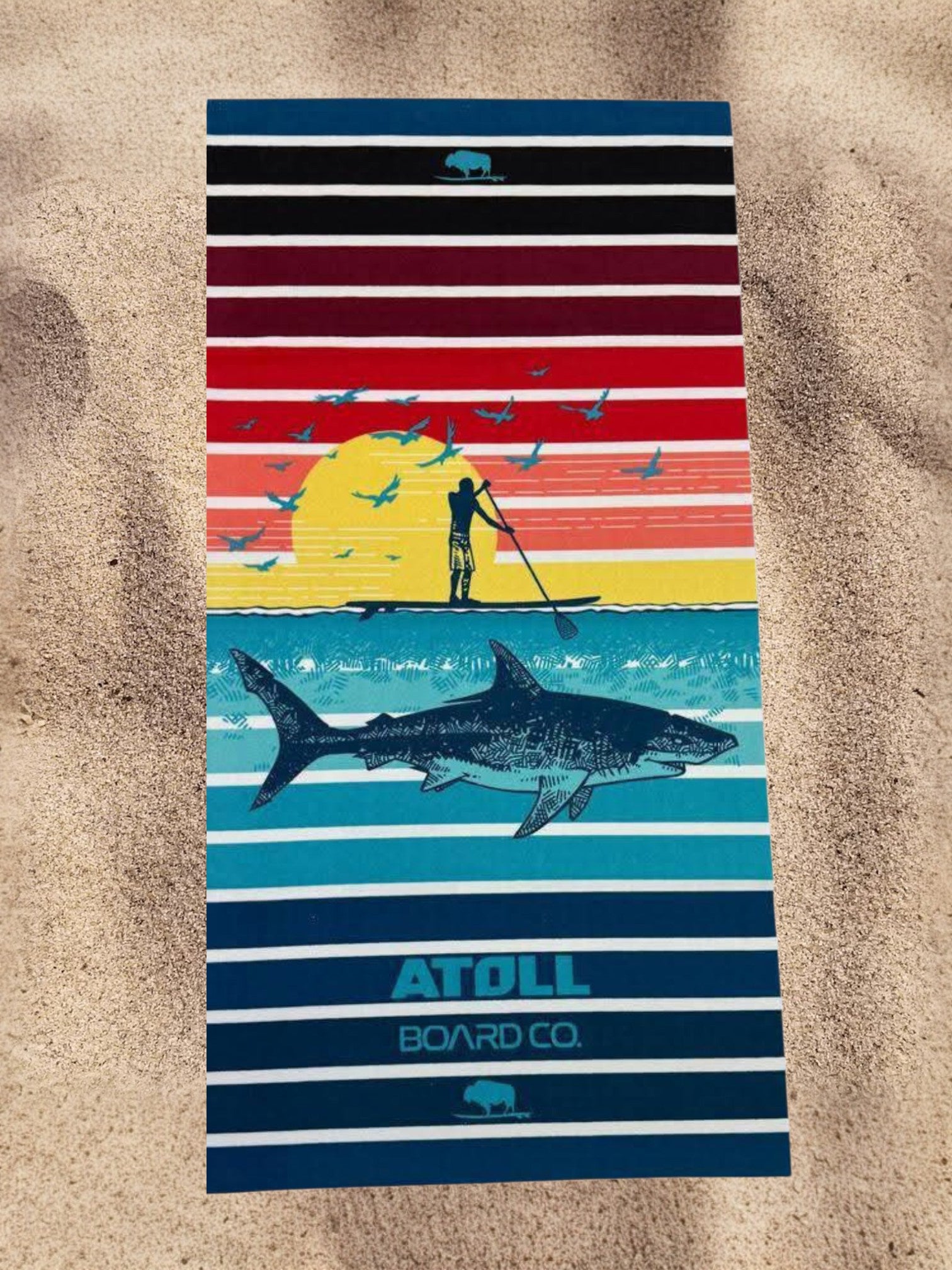 Striped towel with beach scene design and 'Atoll Board Co' branding on sand background
